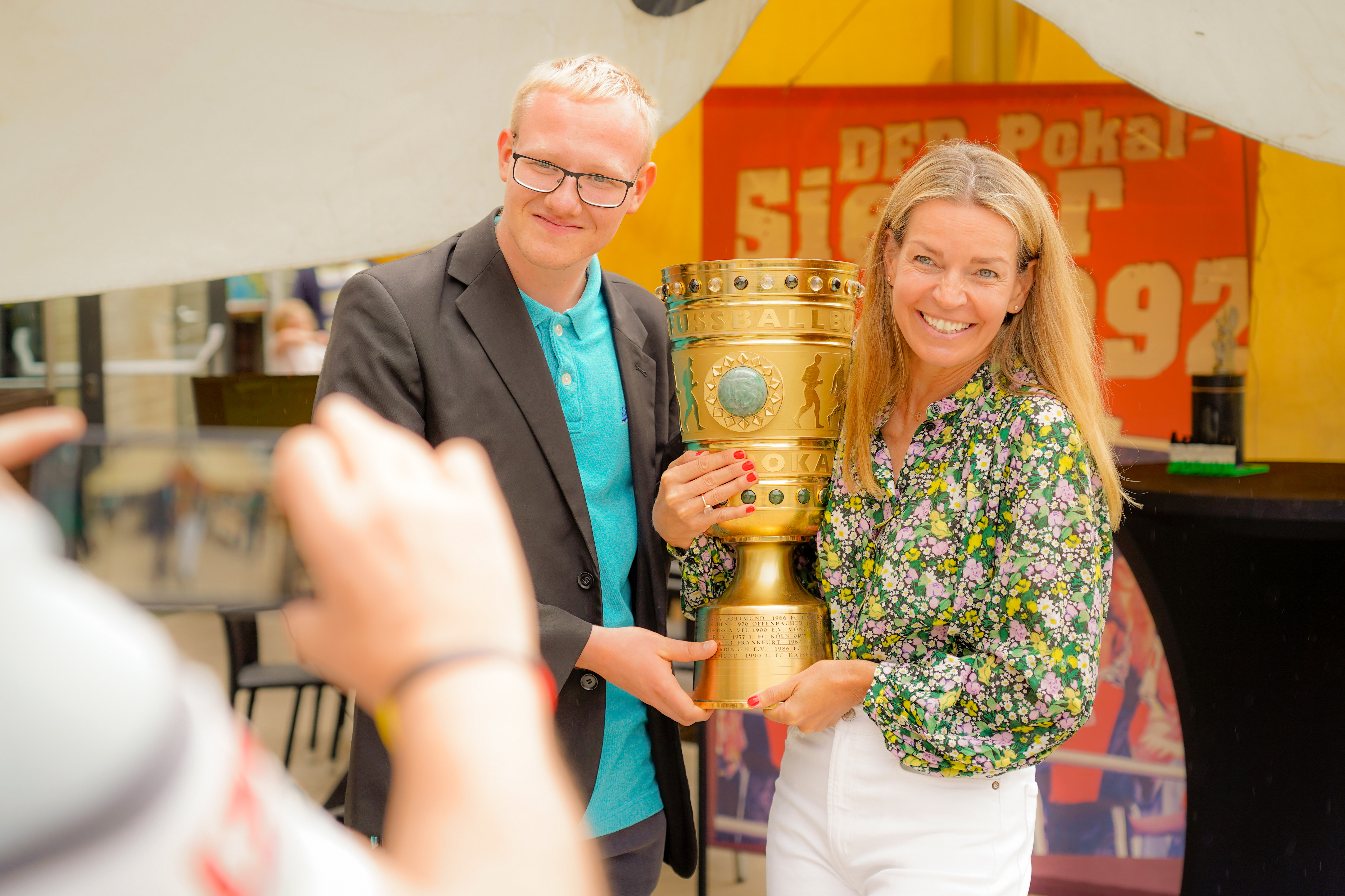 Zwei lächelnde Menschen halten eine große goldene Trophäe zusammen, während jemand unter einem gelben Zelt ein Foto von ihnen macht.
