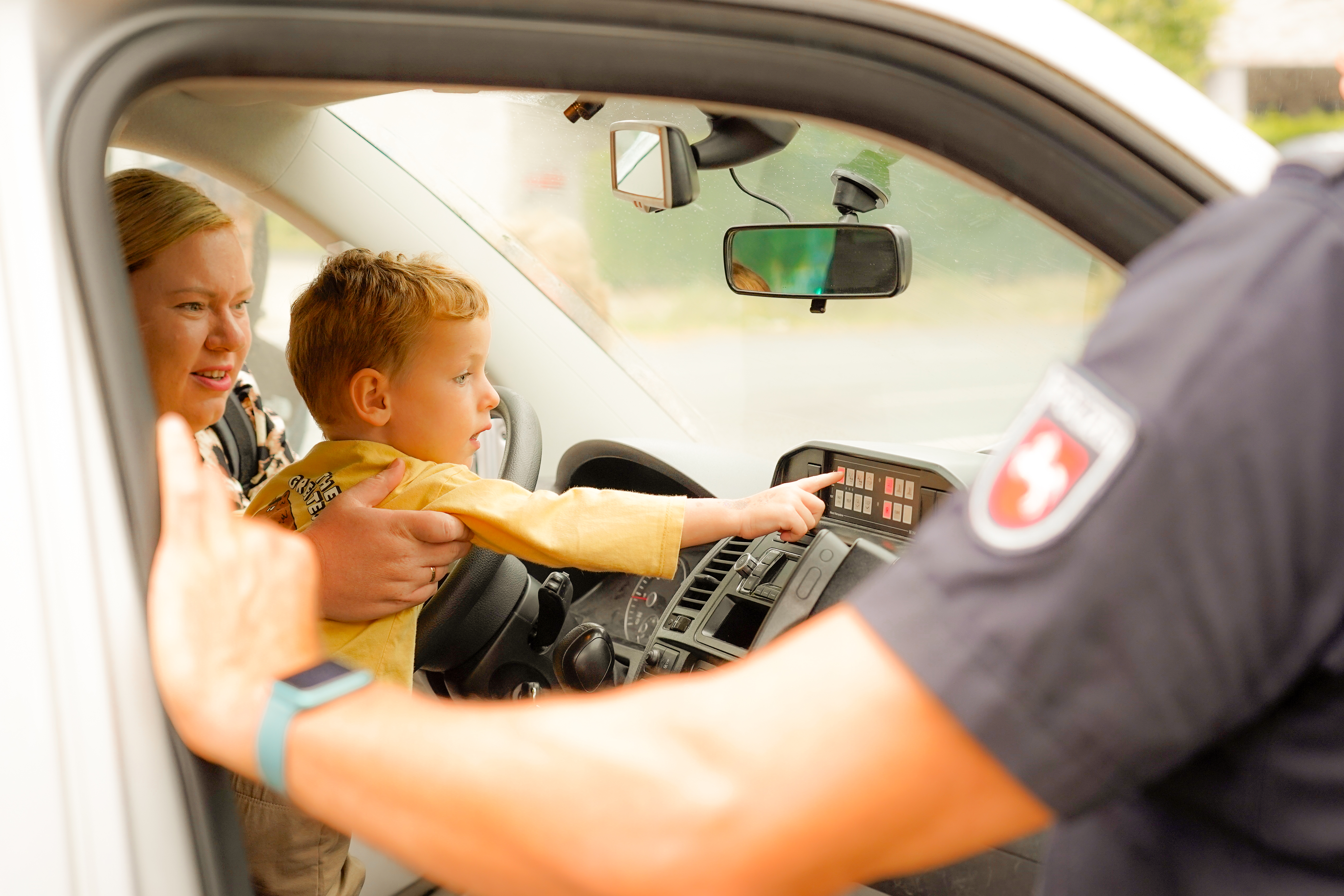Ein Kind sitzt in einem Polizeiauto und zeigt auf das Armaturenbrett, während ein Erwachsener und ein Polizist in der Nähe sind.