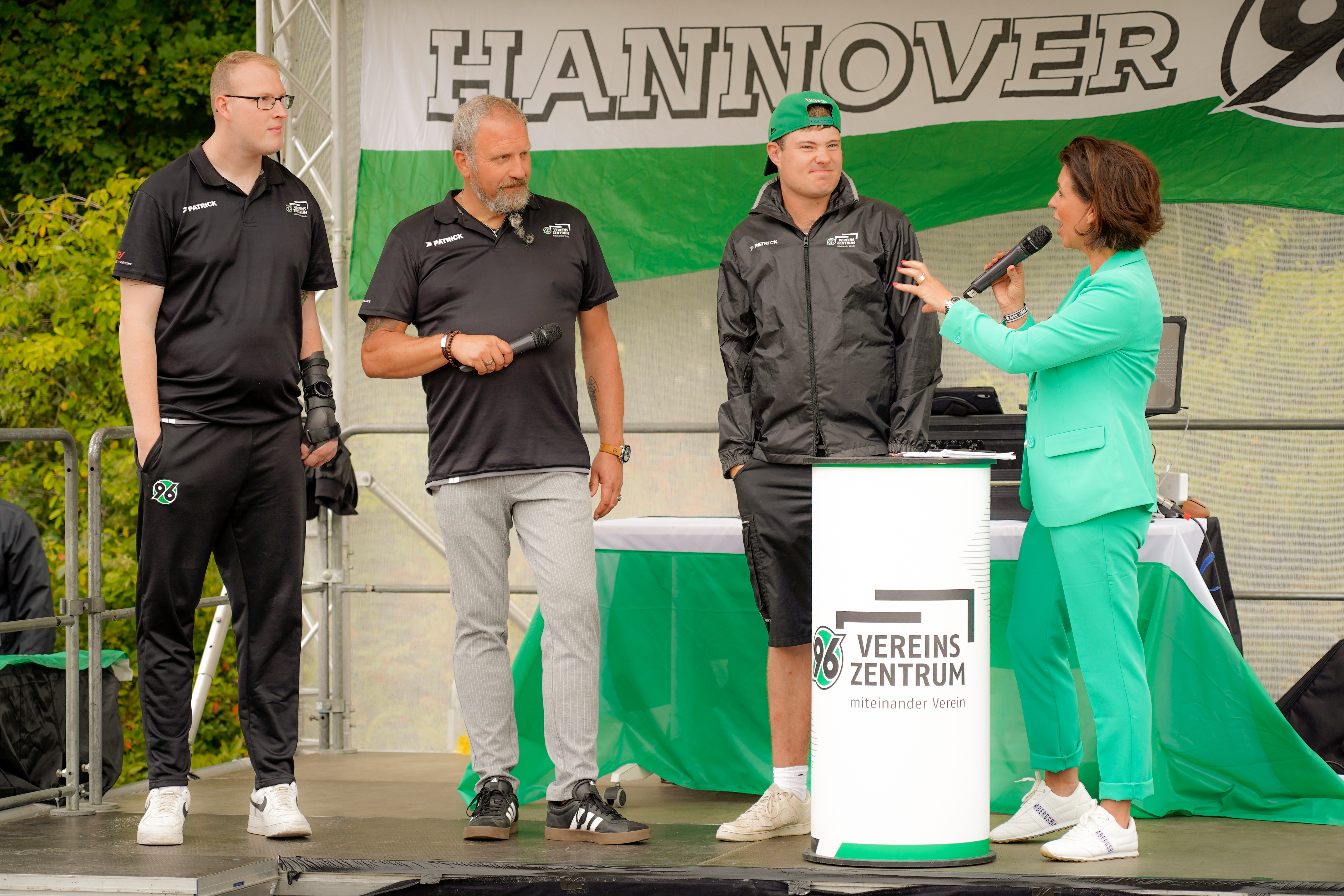 Vier Personen auf der Bühne einer Veranstaltung von Hannover 96, eine Frau in Grün hält ein Mikrofon und interviewt drei Männer.
