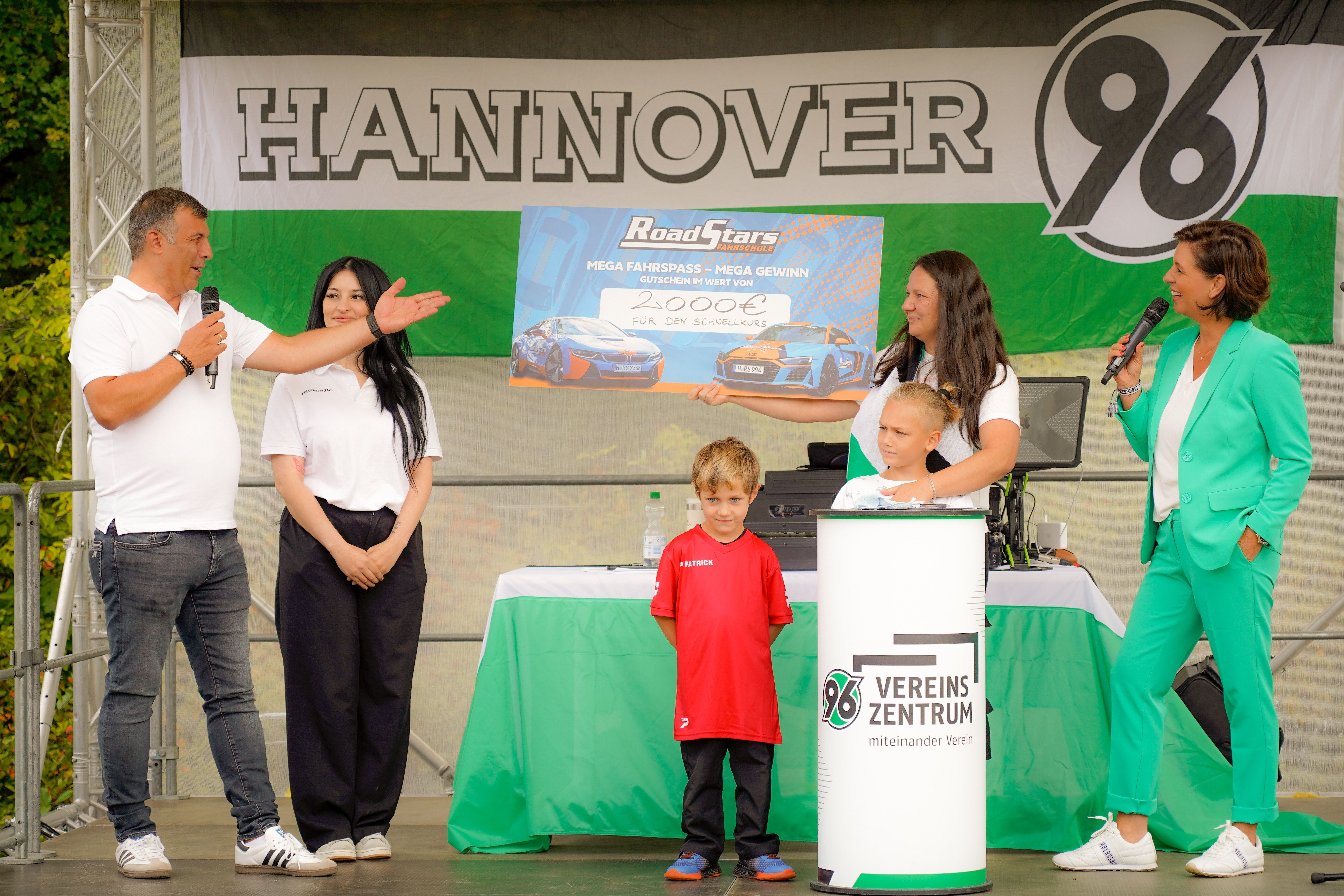 Fünf Personen und ein Kind auf der Bühne mit einem großen Scheck, einer Hannover 96-Fahne und einem Mikrofon; Veranstaltung im Freien.