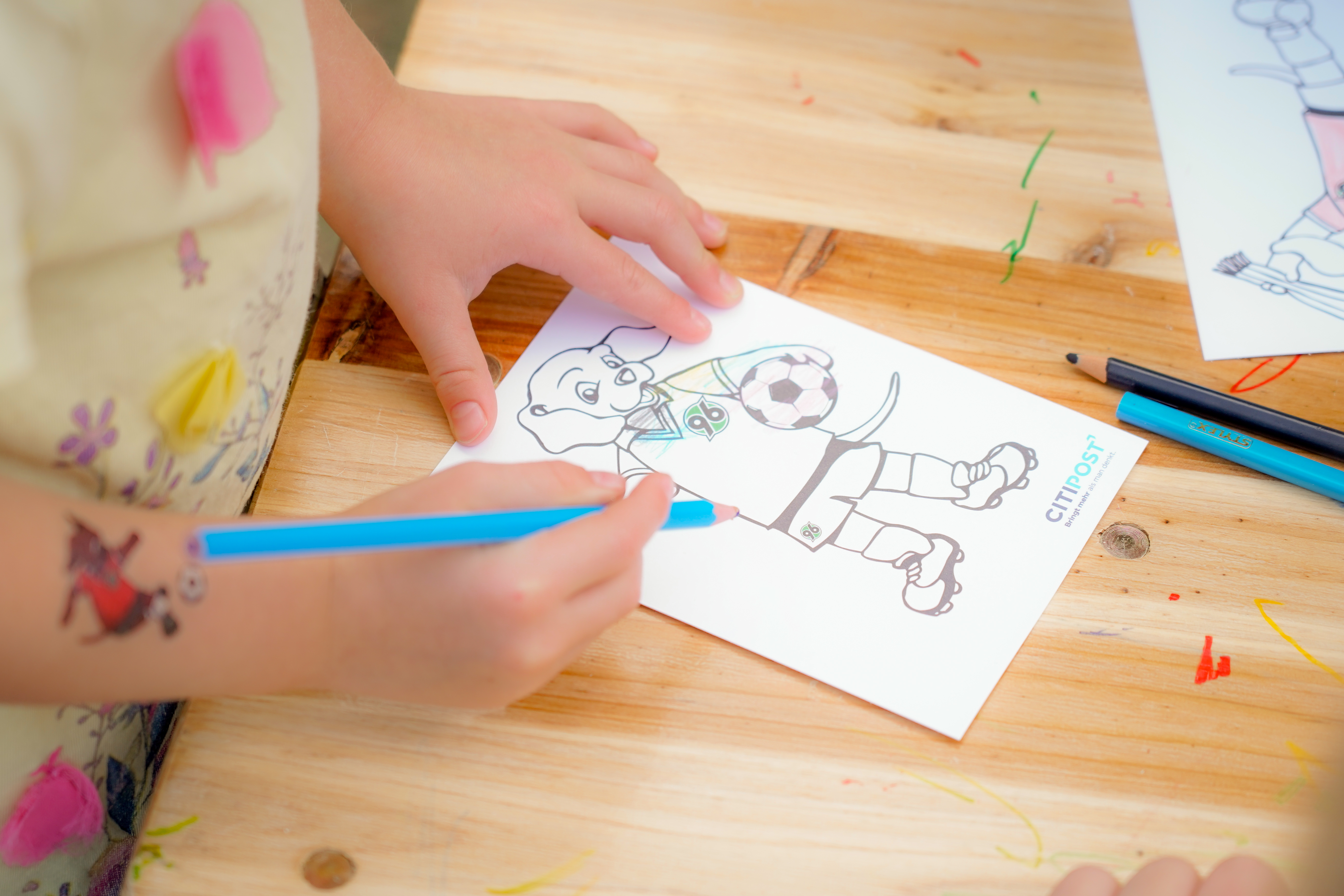 Ein Kind malt mit einem blauen Stift auf einem Holztisch eine Zeichnung zum Thema Fußball.